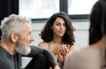 A woman with curly hair sits indoors, holding a cup and smiling, while two other people sit nearby, slightly out of focus. The atmosphere appears relaxed and social.