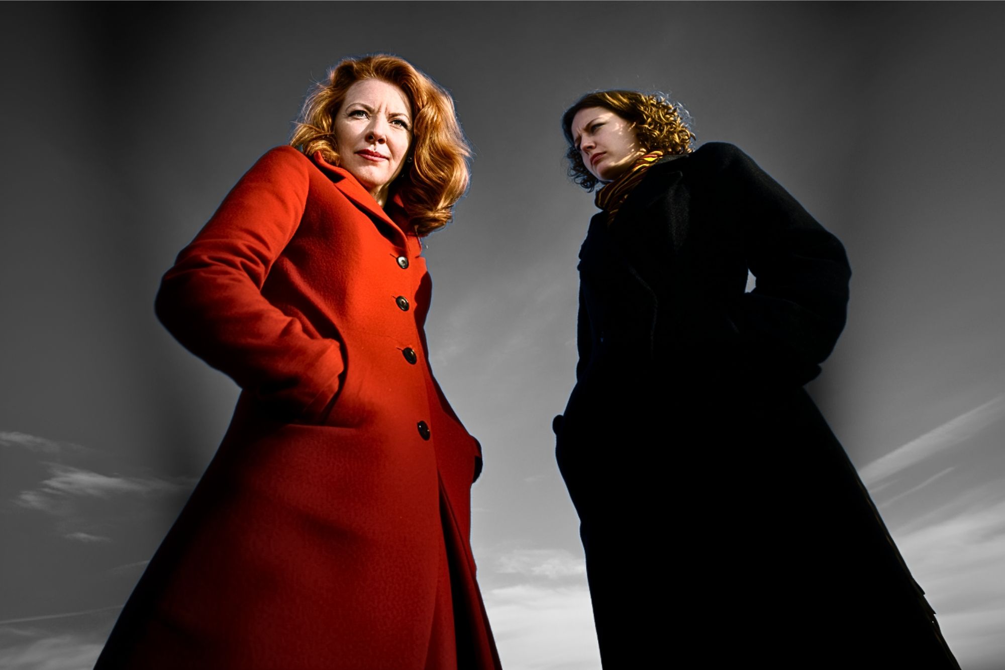 Two women stand outdoors against a dramatic, cloudy sky. One wears a bright red coat and looks confidently at the camera, while the other in a dark coat looks off to the side. The image is taken from a low angle.