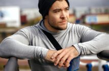 A young man wearing a gray hoodie and black beanie leans on a railing outdoors, looking thoughtfully into the distance. He has a smartphone armband and appears to be taking a break from exercise.