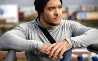 A young man wearing a gray hoodie and black beanie leans on a railing outdoors, looking thoughtfully into the distance. He has a smartphone armband and appears to be taking a break from exercise.