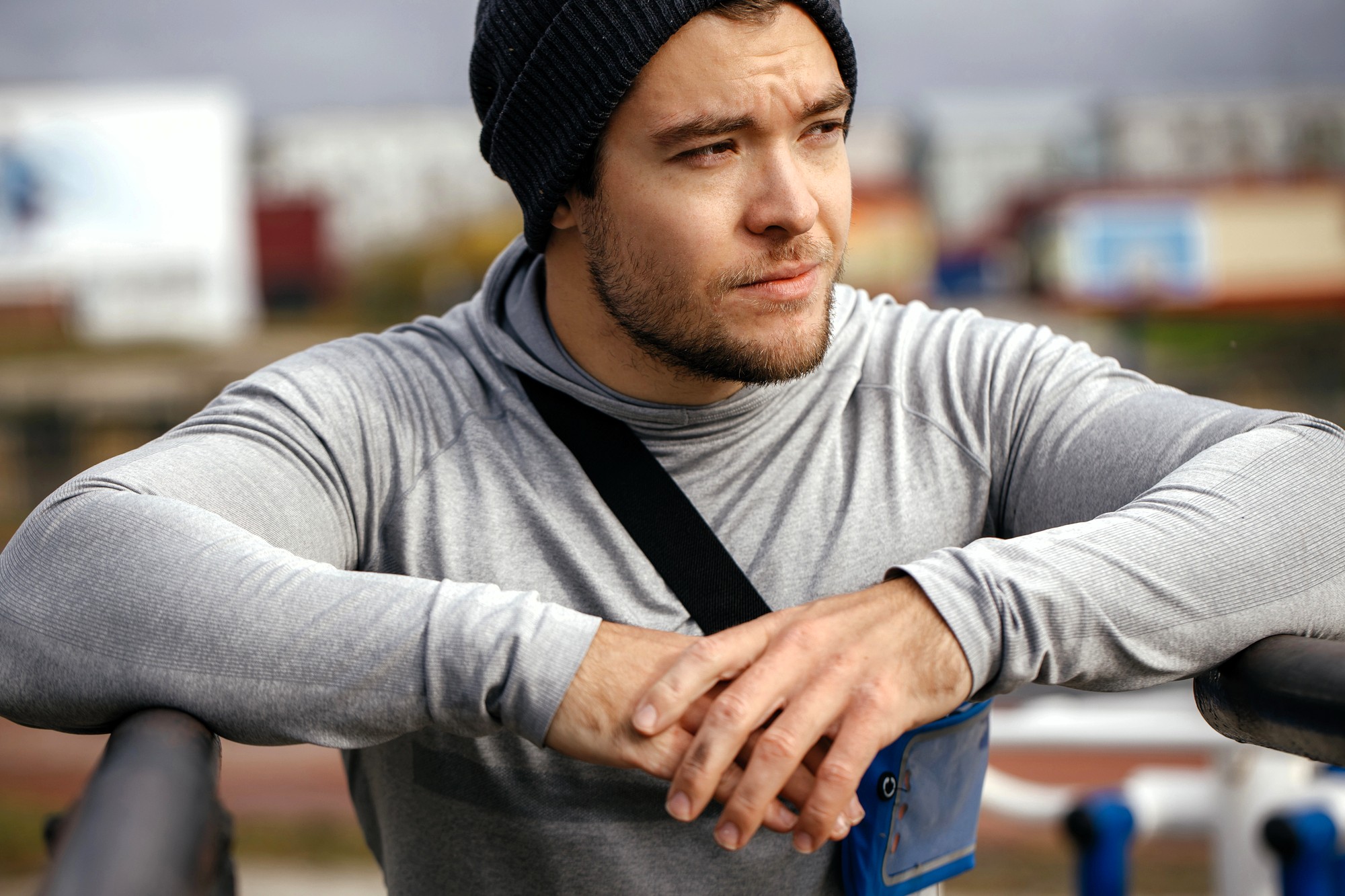 A young man wearing a gray hoodie and black beanie leans on a railing outdoors, looking thoughtfully into the distance. He has a smartphone armband and appears to be taking a break from exercise.