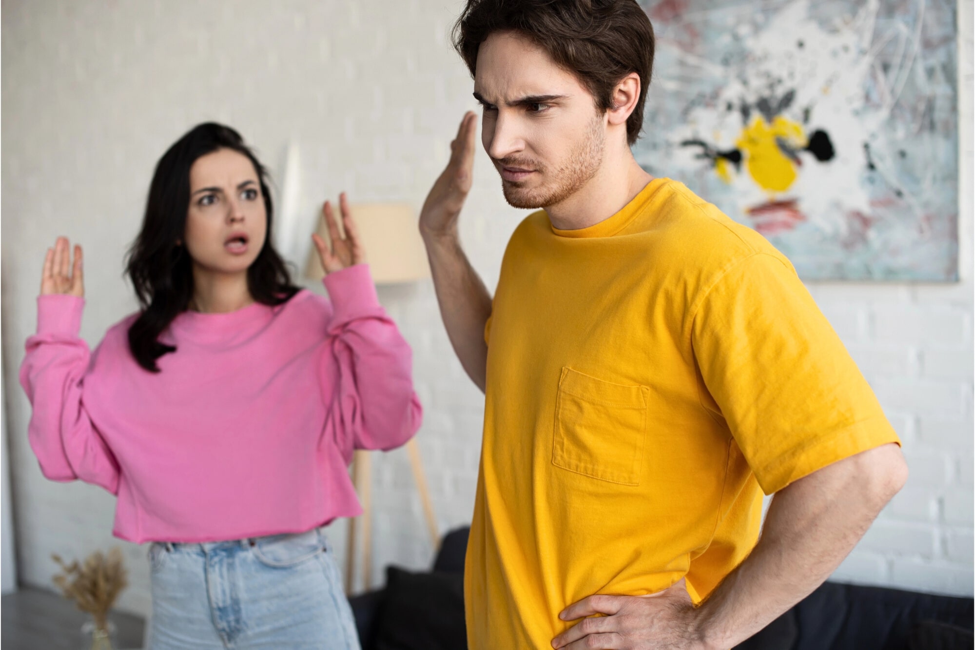 A woman in a pink sweater gestures with raised hands, looking upset, while a man in a yellow shirt turns away with a stern expression, raising his hand as if to stop her.