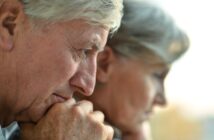 Close-up of an elderly man and woman, both with gray hair, looking pensive and thoughtful. Their faces are partially in profile, with the man in sharp focus and the woman blurred in the background.