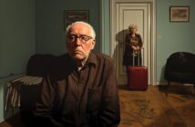 An elderly man sits looking sad in a dimly lit room, while an elderly woman stands in the background holding the handle of a red suitcase near a closed door, suggesting she may be leaving.
