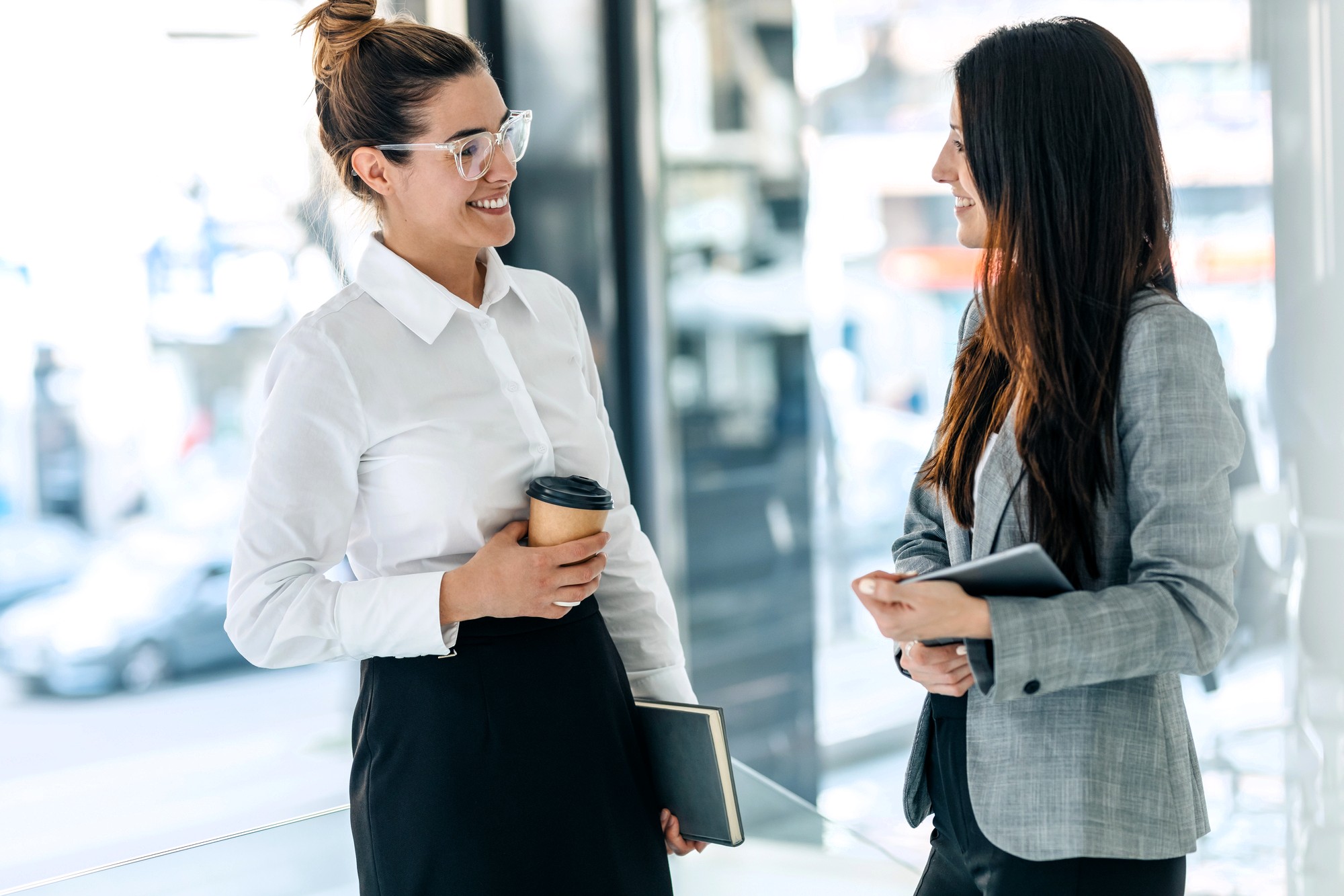 Two women in business attire stand indoors, smiling and talking. One holds a coffee cup and notebook, while the other holds a tablet. Bright, modern office environment with large windows in the background.