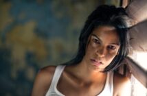 A woman with long dark hair and a white tank top leans against a wooden window shutter, looking directly at the camera with a serious expression. The background is dim and out of focus.
