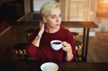 A person with short blond hair wearing a red sweater sits at a table in a café, holding a cup of coffee and looking thoughtfully into the distance. An empty saucer is on the table in front of them.