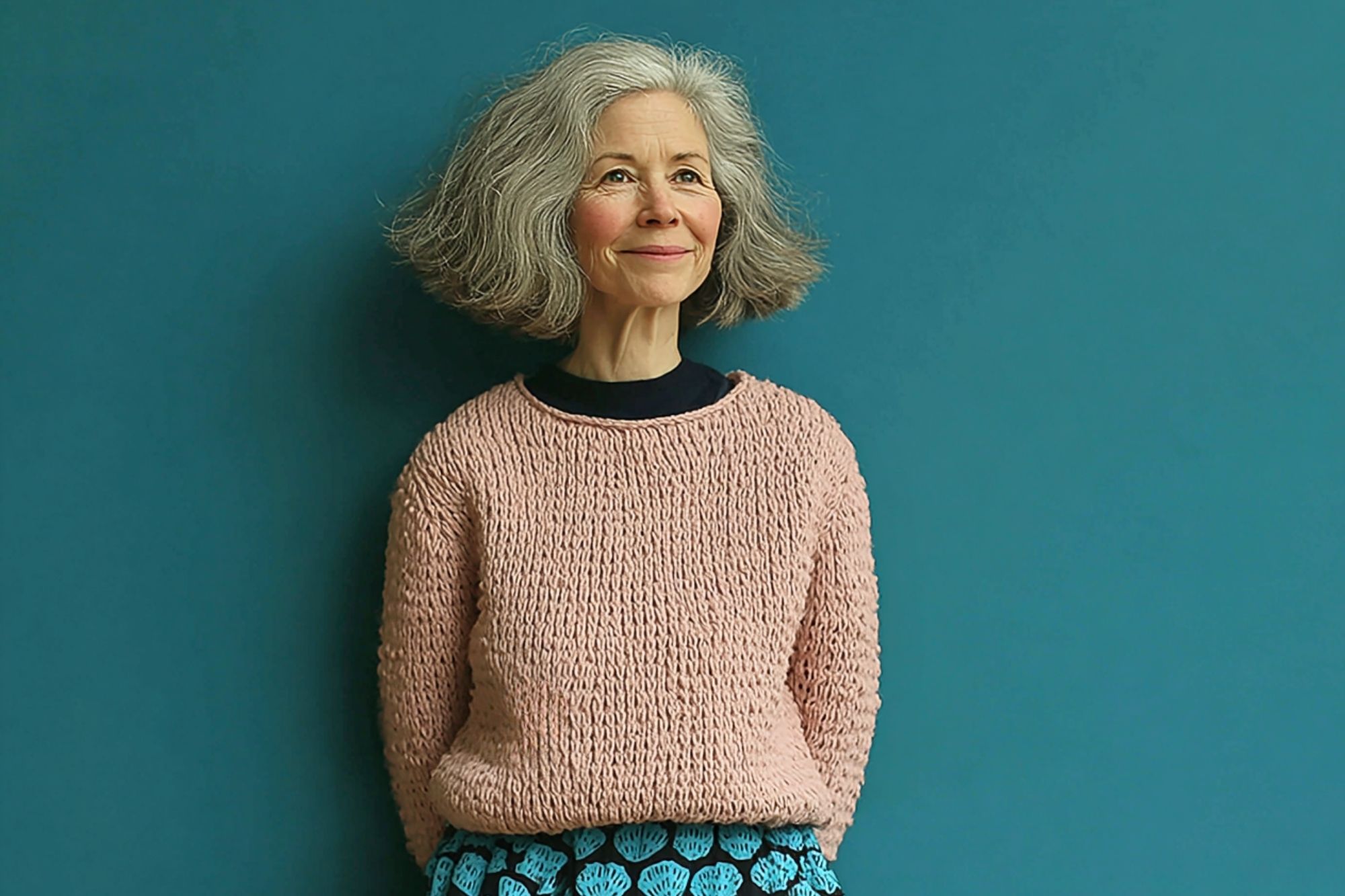 An older woman with gray, shoulder-length hair smiles gently while standing against a teal wall. She wears a pale pink sweater and a skirt with a blue and black heart pattern.