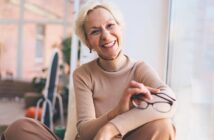 An older woman with short blonde hair, wearing a beige turtleneck and pants, sits by a window smiling warmly while holding a pair of glasses in her hand. Soft natural light fills the room.