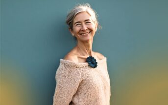 An older woman with gray hair smiles warmly, wearing an off-the-shoulder beige knit sweater and a dark blue flower pin. She stands against a plain teal background.