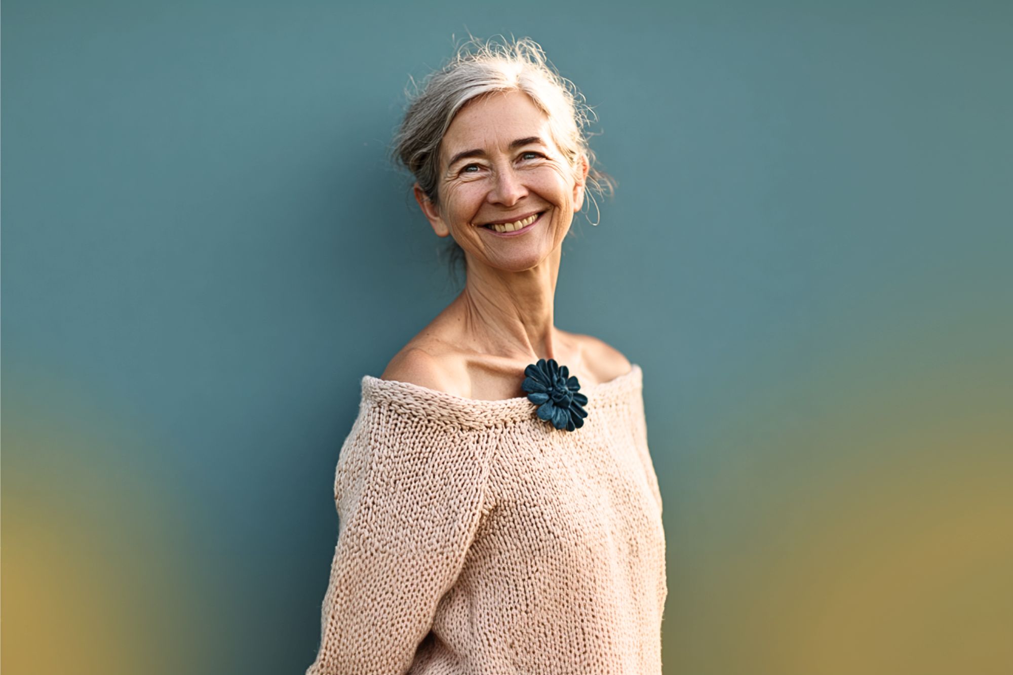 An older woman with gray hair smiles warmly, wearing an off-the-shoulder beige knit sweater and a dark blue flower pin. She stands against a plain teal background.