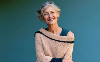 Smiling older woman with gray hair wears a beige sweater with dark trim, standing against a solid teal background, arms gently crossed.