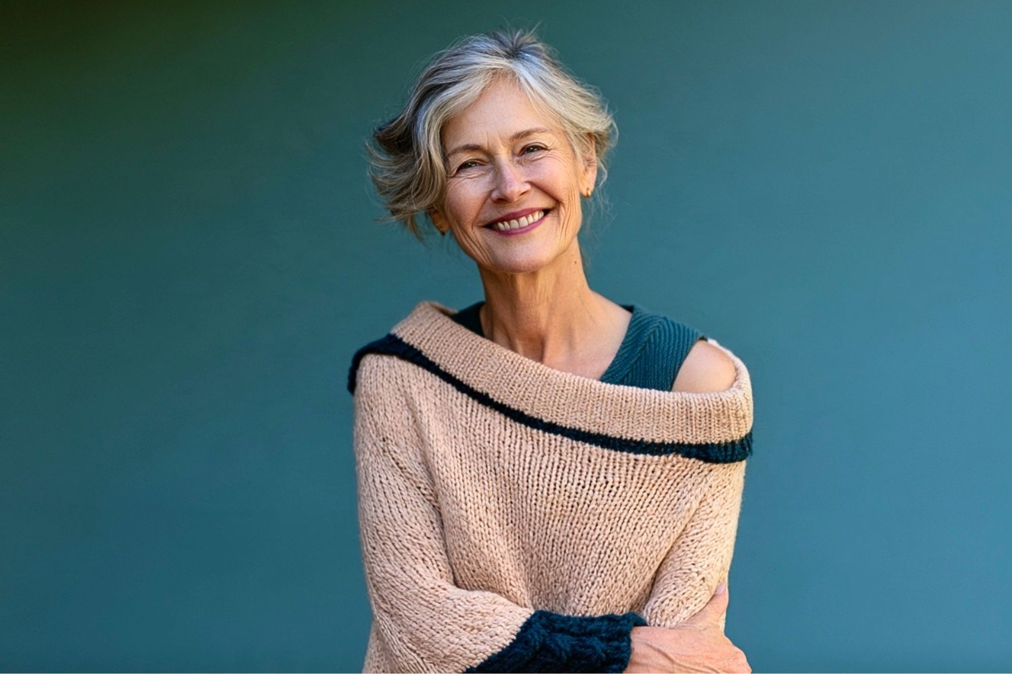 Smiling older woman with gray hair wears a beige sweater with dark trim, standing against a solid teal background, arms gently crossed.