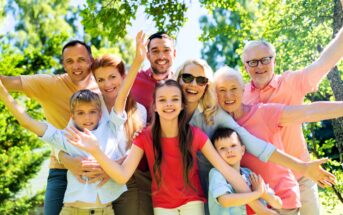 A large, happy family of multiple generations stands close together outdoors, smiling and posing with arms outstretched on a sunny day surrounded by green trees.