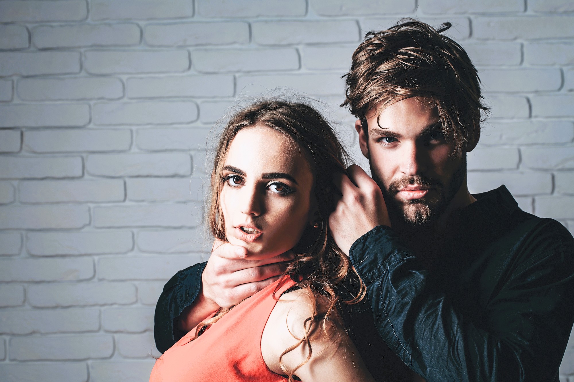 A woman in an orange top stands in front of a man in a black shirt. The man is gripping her chin and neck with one hand. Both have serious expressions and stand against a white brick wall.