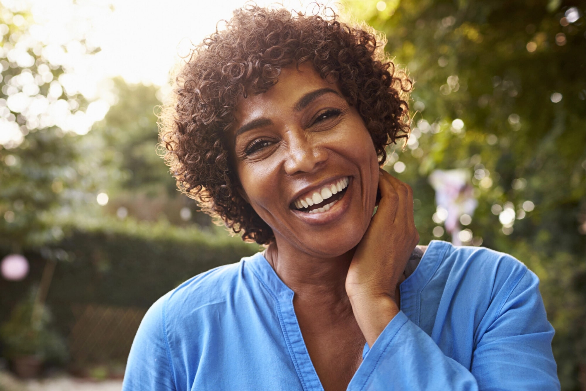 A woman with short curly hair wearing a blue top smiles warmly at the camera outdoors, with one hand gently touching her neck. The background is blurred with greenery and sunlight.