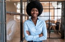 A woman with a natural afro hairstyle, wearing a light blue button-up shirt, stands confidently with arms crossed and smiles in a modern office with glass walls and wooden floors.
