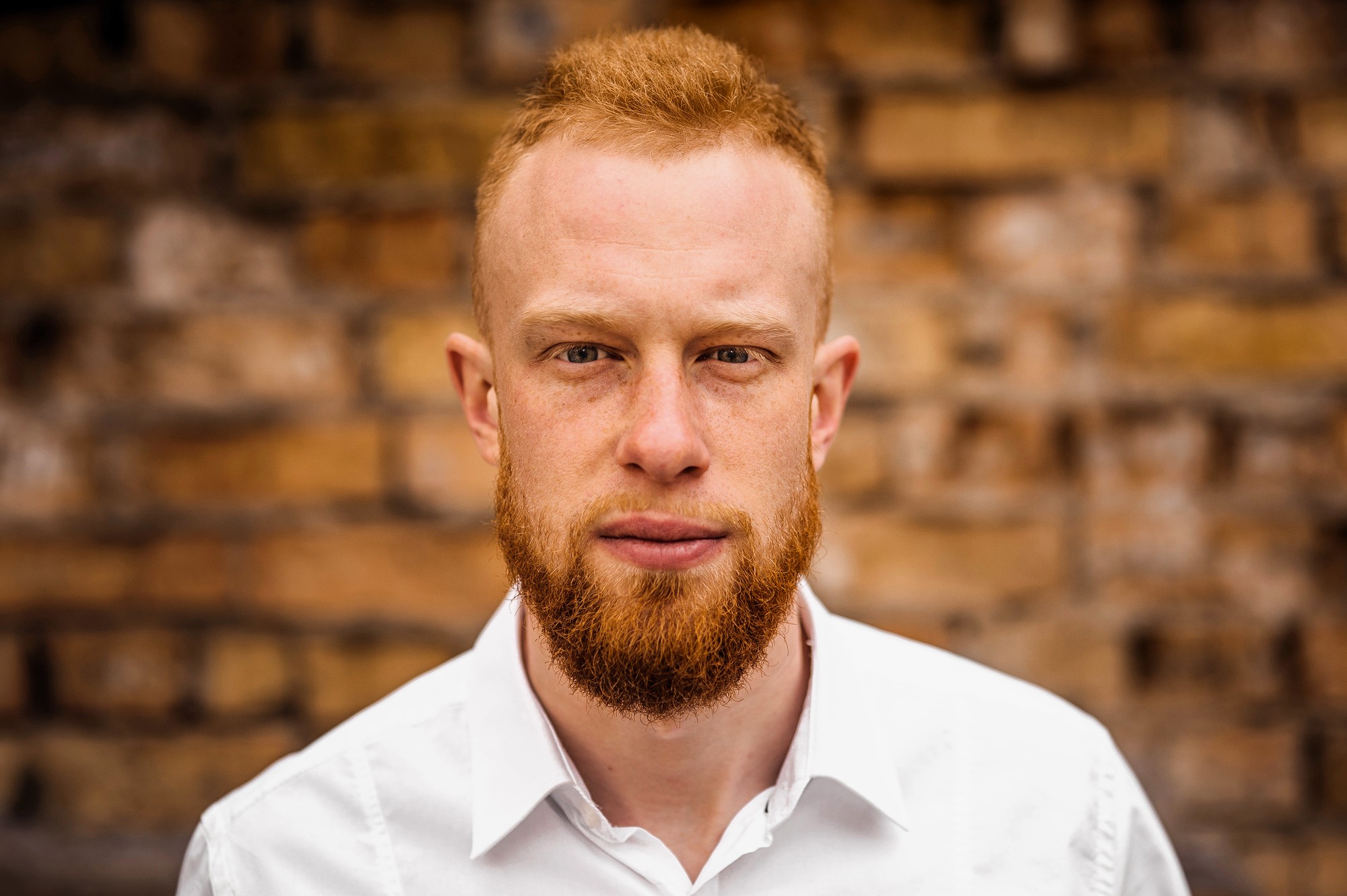 A man with short red hair and a full red beard, wearing a white collared shirt, stands in front of a rustic brick wall and looks directly at the camera with a neutral expression.
