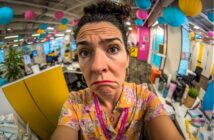 A woman with curly hair and a floral shirt makes an exaggerated, confused face in a colorful, open office decorated with hanging paper lanterns. The photo is taken with a fisheye lens, distorting the scene.