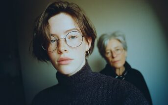 A young person with short hair and round glasses takes a selfie, wearing a dark sweater, while an older person with gray hair and glasses stands blurred in the background.