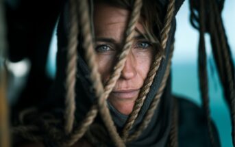 A person with blue eyes and light brown hair peers through thick, weathered ropes. Their face is framed by the ropes and a hood, with soft natural light highlighting their freckles and focused expression.