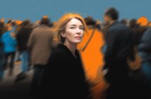 A woman with reddish hair looks up pensively, standing still amid a blurred crowd of people moving around her. The background is dark and blue, with orange highlights, making her stand out in sharp focus.