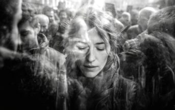 A black and white photo shows a woman with closed eyes standing amidst a crowd. The image is double-exposed, creating a dreamy, reflective effect with her face partially overlaid by other people.