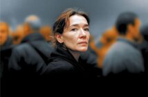 A woman with short brown hair and a serious expression stands in focus among a blurred crowd of people in dark clothing, with an orange and gray background.