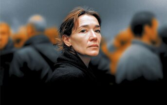 A woman with short brown hair and a serious expression stands in focus among a blurred crowd of people in dark clothing, with an orange and gray background.