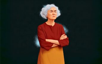An older woman with short white hair stands with arms crossed, wearing a red top and orange skirt, looking serious against a dark background.