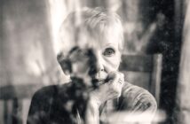 An elderly person with short hair gazes thoughtfully out a window, resting their chin on their hand. The image is black and white with soft focus and reflections on the glass.