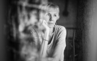 Black and white photo of an older woman with short hair, resting her chin on her hand and gazing thoughtfully out of a window. She appears pensive and lost in thought.