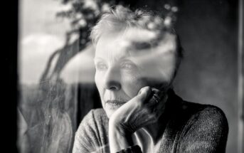 An elderly woman gazes thoughtfully out a window, resting her chin on her hand. The black and white photo captures her reflection and a blurry outdoor scene in the glass.