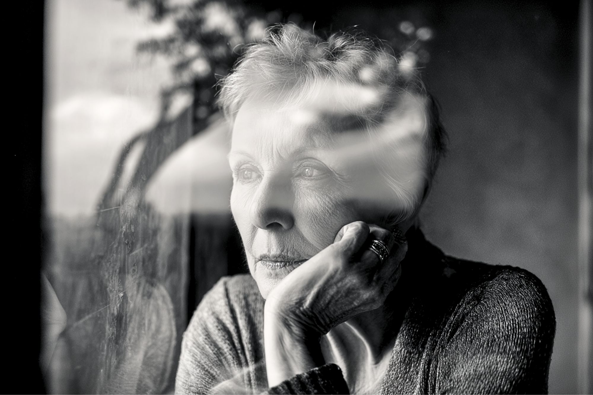 An elderly woman gazes thoughtfully out a window, resting her chin on her hand. The black and white photo captures her reflection and a blurry outdoor scene in the glass.