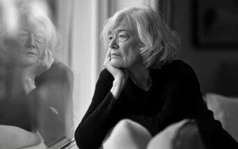 An older woman with gray hair resting her chin on her hand, looking out a window.