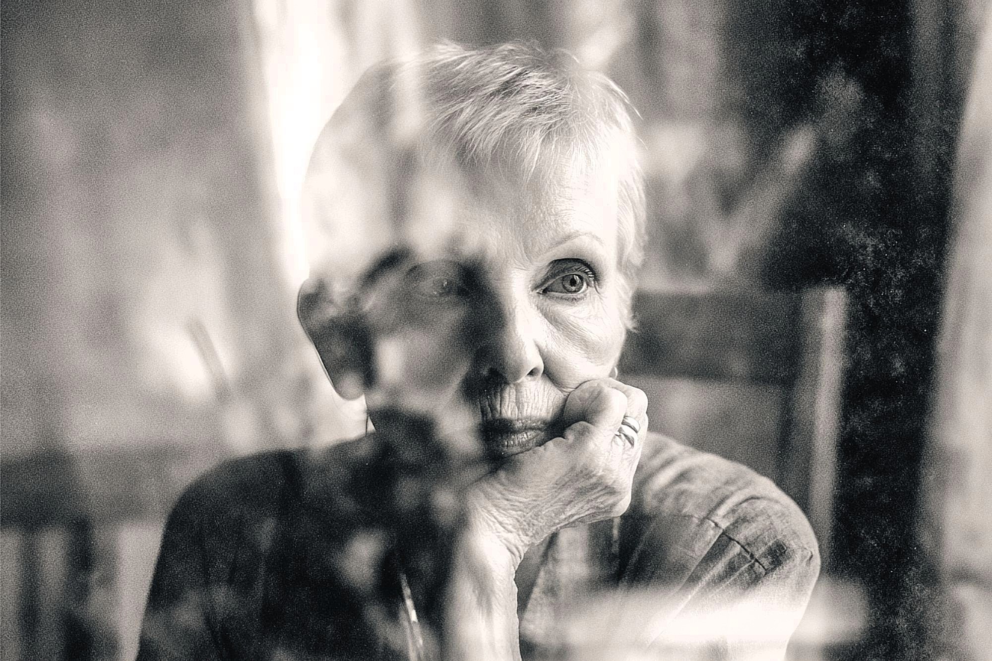 An elderly person with short hair gazes thoughtfully out a window, resting their chin on their hand. The image is black and white with soft focus and reflections on the glass.