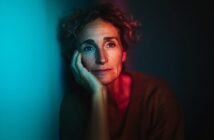 A woman with curly hair rests her face on her hand, gazing thoughtfully into the distance, illuminated by contrasting blue and red light in a dark setting.