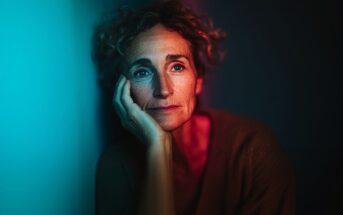 A woman with curly hair rests her face on her hand, gazing thoughtfully into the distance, illuminated by contrasting blue and red light in a dark setting.
