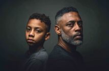 A boy and a man with similar features sit back-to-back, both looking seriously at the camera against a dark background, highlighting their expressions and resemblance.