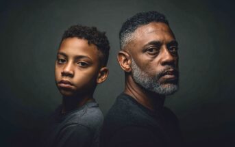 A boy and a man with similar features sit back-to-back, both looking seriously at the camera against a dark background, highlighting their expressions and resemblance.