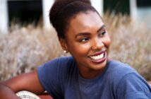 A young woman with dark skin and natural hair smiles brightly while looking to the side. She is wearing a blue shirt and stud earrings, sitting outdoors in front of blurred plants.