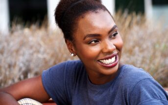A young woman with dark skin and natural hair smiles brightly while looking to the side. She is wearing a blue shirt and stud earrings, sitting outdoors in front of blurred plants.