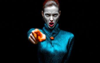 A woman with slicked-back hair and hoop earrings wears a textured blue sweater with a large button. She points directly at the camera, staring intensely with an open mouth against a black background.