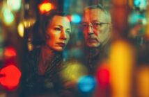 A middle-aged woman and man sit close together, gazing pensively to the side in a dim, colorful, and moody setting with blurred, vibrant lights in the foreground and background.