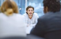 A young man with long hair and a white shirt sits at a table, facing two people whose backs are to the camera, in a bright office setting. He appears to be speaking or answering questions.