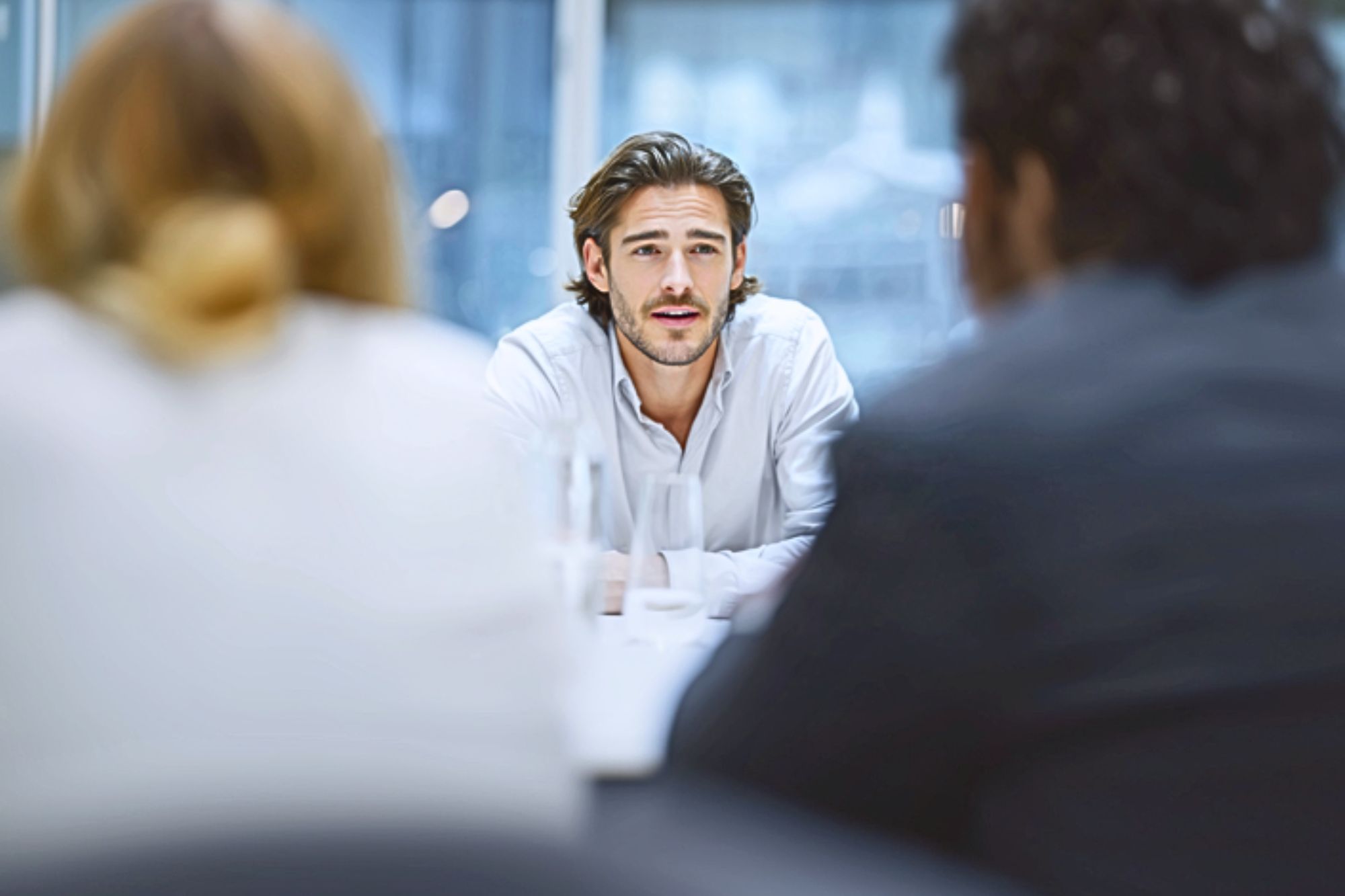 A young man with long hair and a white shirt sits at a table, facing two people whose backs are to the camera, in a bright office setting. He appears to be speaking or answering questions.