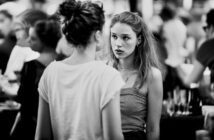 Two young women have a serious conversation in a crowded, blurred background. One faces the camera with a concerned expression, while the other has her back to the camera. The image is in black and white.