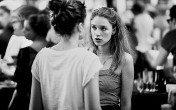 Two young women have a serious conversation in a crowded, blurred background. One faces the camera with a concerned expression, while the other has her back to the camera. The image is in black and white.