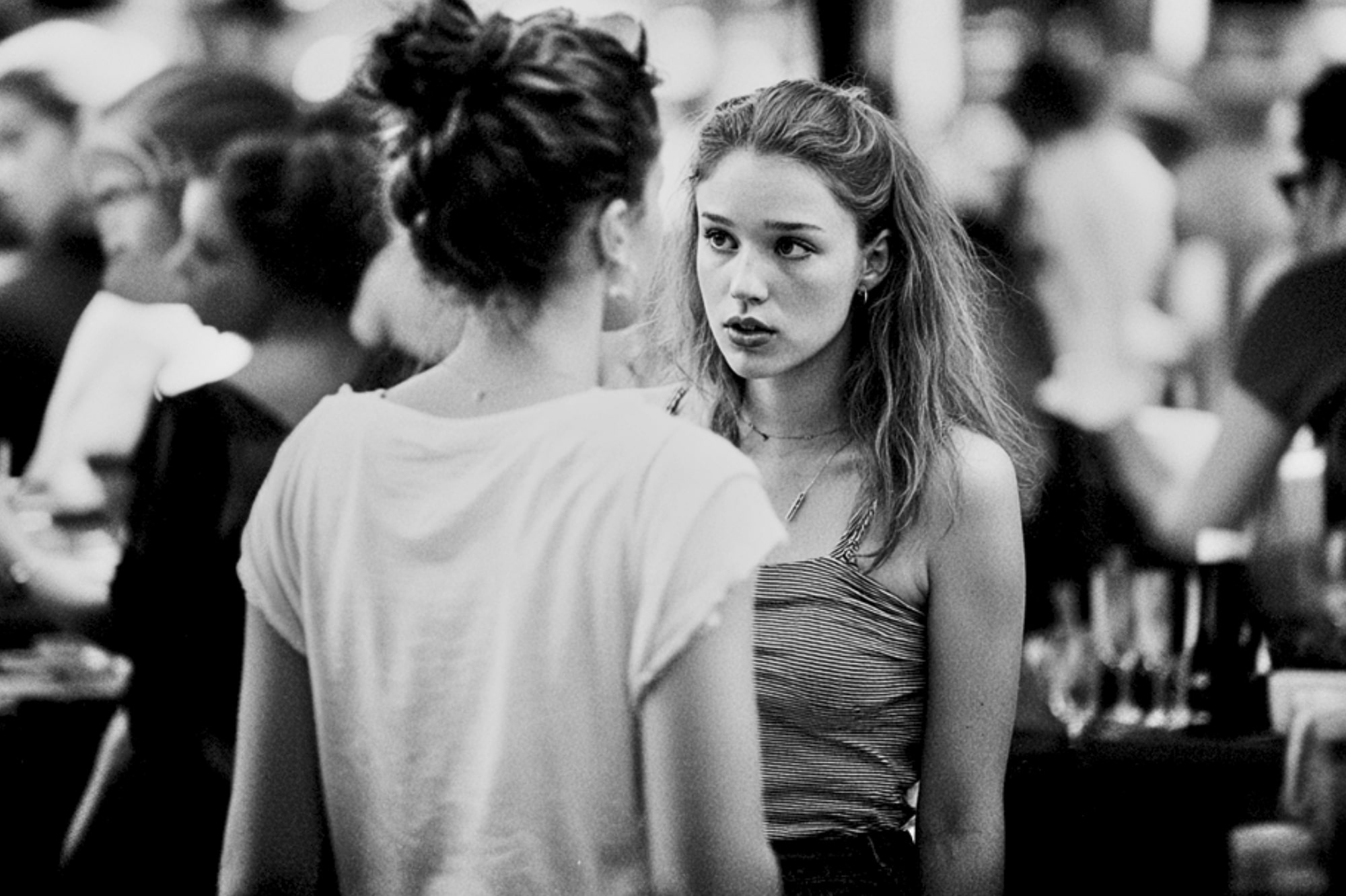 Two young women have a serious conversation in a crowded, blurred background. One faces the camera with a concerned expression, while the other has her back to the camera. The image is in black and white.
