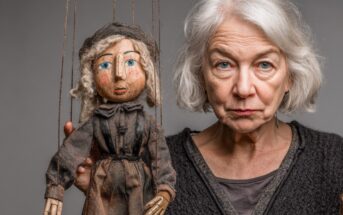 An older woman with gray hair stands next to a marionette puppet that resembles her, both wearing serious expressions. The puppet is dressed in old-fashioned clothes and has blue eyes and curly hair.